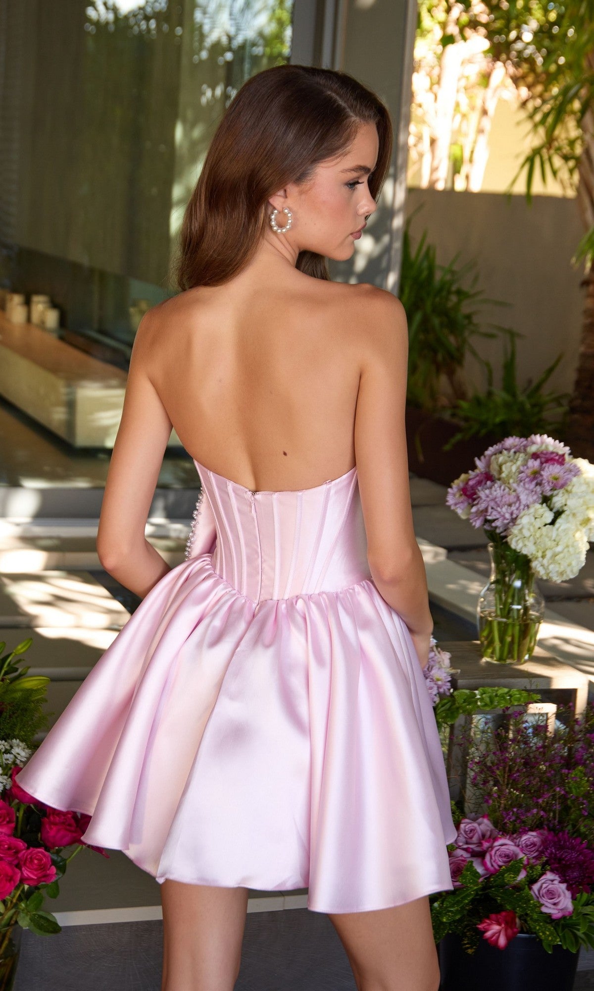A woman stands with her back to the camera among flower arrangements, wearing the Ava Presley 47298 short homecoming dress featuring a strapless satin design and boned corset bodice.